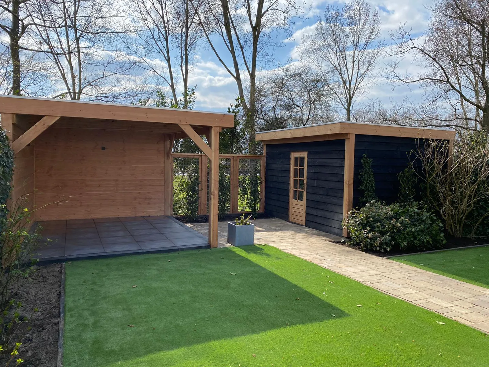 Houten carport naast een tuinhuis met kunstgras